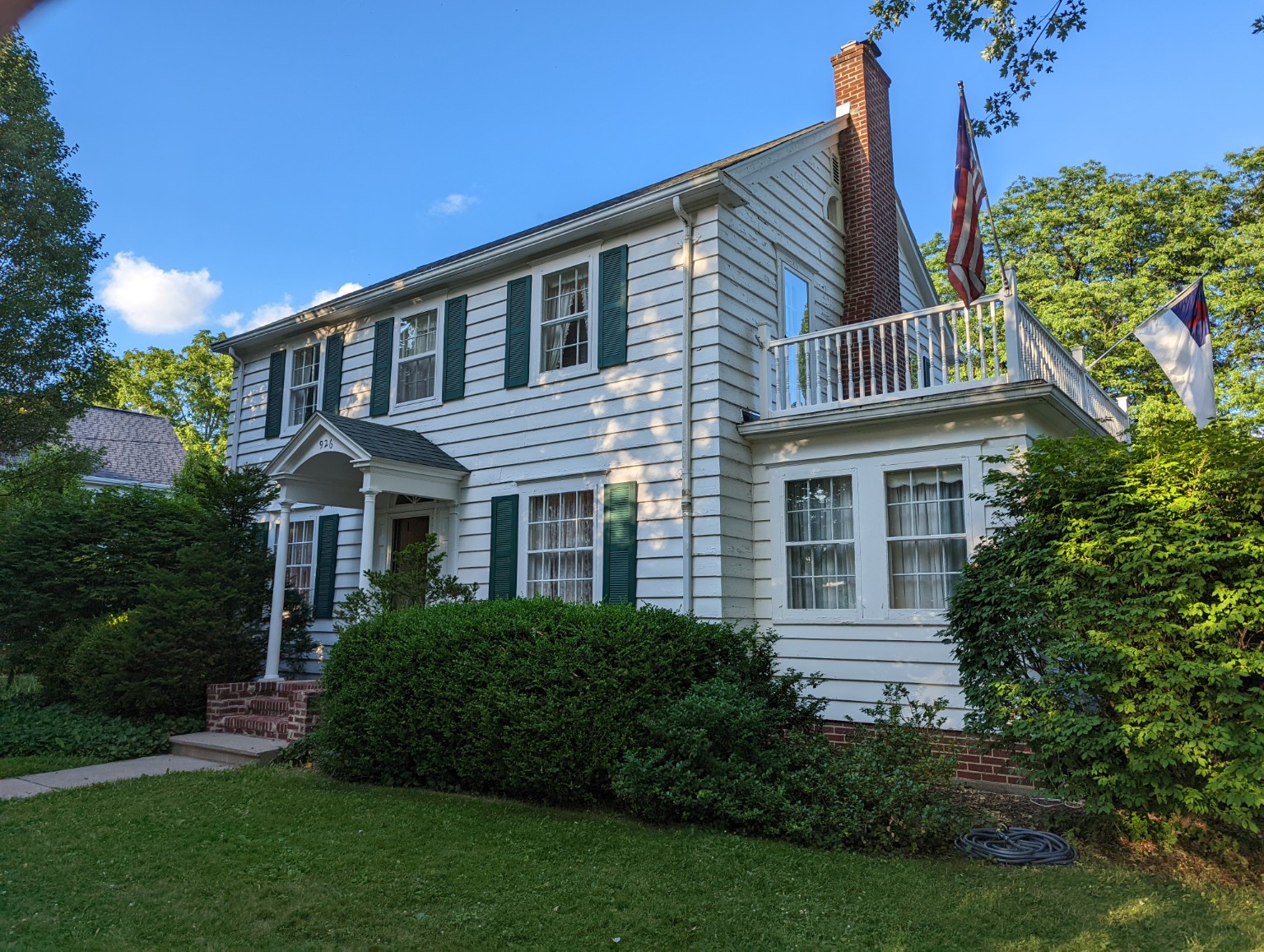 926 N. Washington Street in July 2022, showing white siding, green shutters, a columned entrance and brick chimney.